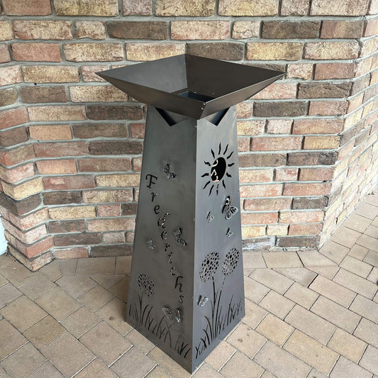 Ansicht von oben auf eine Viereckige Blumensäule aus Metall in Silber. Auf der Linkenseite der Säule steht ein Name auf den anderen Seiten sind Blumen und Schmetterlinge