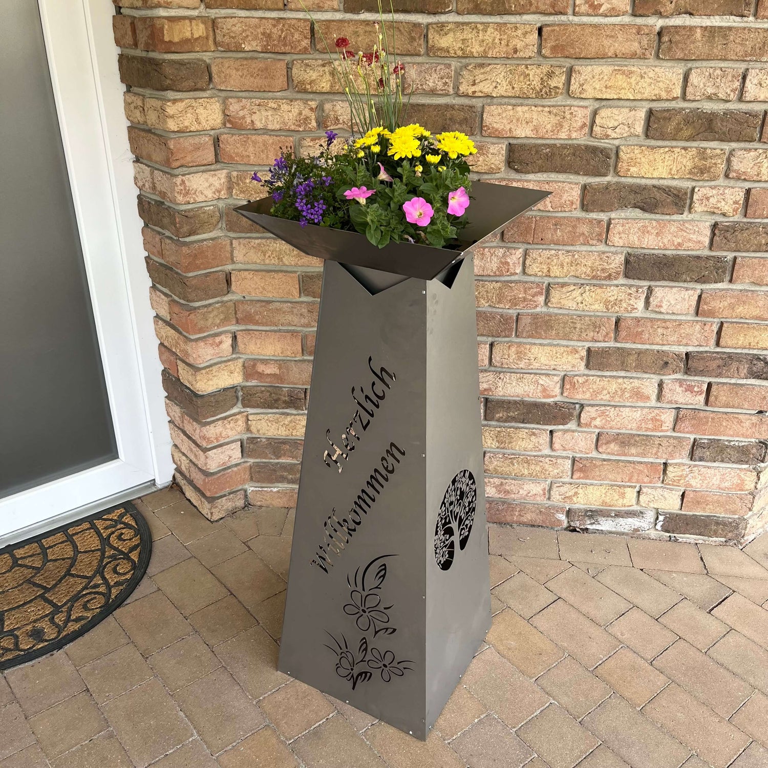 Graue Rechteckige Säule mit großer eckiger Schale mit pinken, violetten, roten und gelben Blumen vor einer Steinmauer.