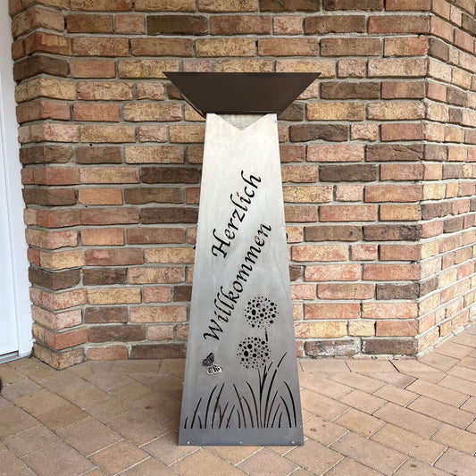 Blumensäule in Silber vor einer Hauswand, in der Blumensäule steht Herzlich Willkommen und im unteren Bereich sind ausgeschnittene Blumen und ein Schmetterling.