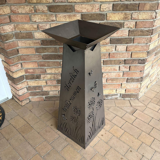 Rechteckige hoche Säule in grau, nach oben hin zulaufend, Rechteckige Pflanzenschale oben drauf, vor Hausmauer. Auf der Säule steht Herzlich Willkommen mit Blumen und Schmetterlingen.
