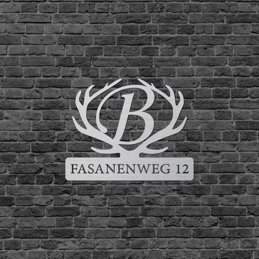 Eine Wanddekoration mit einem Hirschgeweih. In der Mitte des Hirschgeweihs befindet sich ein großes B und da drunter steht der Straßenname und die Nummer. Das Schild hängt an einer Hauswand.