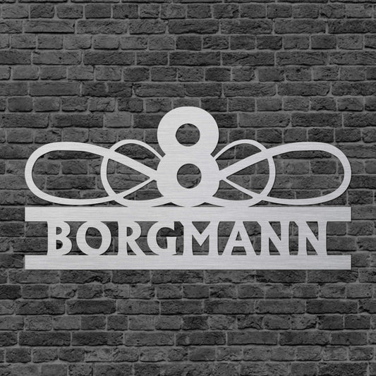 Eine Wanddekoration, wo der Familienname zwischen zwei waagerechten Balken steht und obendrauf befindet sich ein Stahlgeflecht mit einer Zahl in der Mitte. Schild hängt an einer Hauswand.