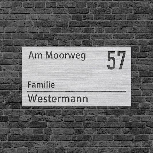 Eine Wanddekoration in Form eines rechteckigen Schilds. an der oberen Kante steht der Straßenname links und rechts die vergrößerte Hausnummer. An der unteren Kante steht das Wort Familie und dann darunter der Familienname. Das Schild hängt an einer Hauswand.