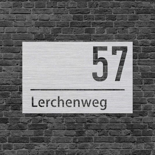 Eine Wanddekoration. In der Mitte ist eine Linie. Oberhalb steht in der rechten Ecke die Hausnummer und unterhalb steht der Straßenname mittig. Das Schild hängt an einer Hauswand.