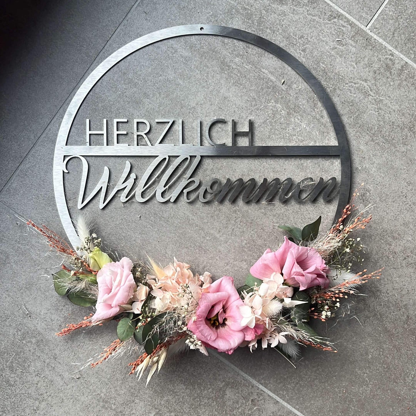 Ein runder Türkranz aus silberfarbenem Edelstahl mit dem Schriftzug ‚Herzlich Willkommen‘. Der untere Teil ist beispielhaft mit rosa Trockenblumen und Gräsern dekoriert. Die Dekoration gehört nicht zum Lieferumfang. Der Kranz liegt auf einem grauen Fliesenboden.