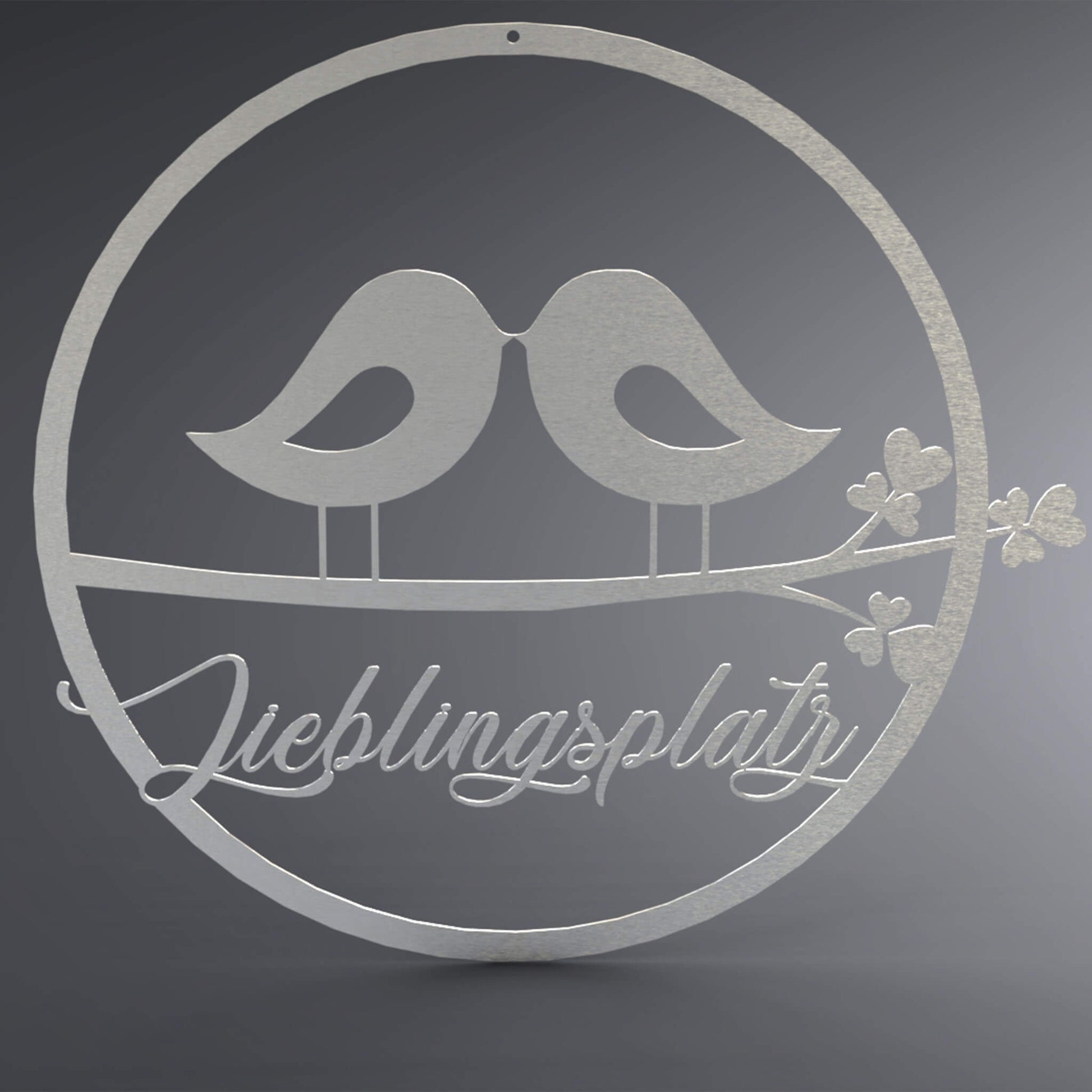 Dünner, ausgehöhlter Kreis. In der Mitte stehen zwei Vögel zueinander gewandt, Schnabel an Schnabel, auf einem Ast. Da drunter steht: "Lieblingsplatz" verschnörkelt.