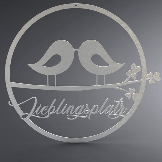 Dünner, ausgehöhlter Kreis. In der Mitte stehen zwei Vögel zueinander gewandt, Schnabel an Schnabel, auf einem Ast. Da drunter steht: "Lieblingsplatz" verschnörkelt.
