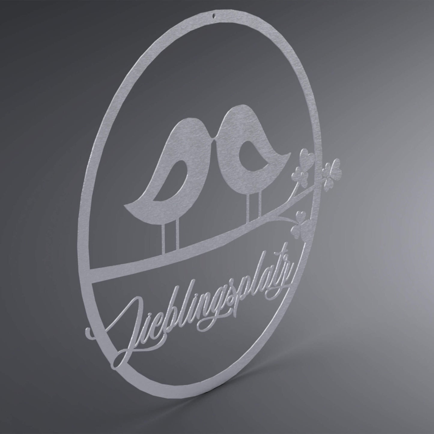 Dünner, ausgehöhlter Kreis. In der Mitte stehen zwei Vögel zueinander gewandt, Schnabel an Schnabel, auf einem Ast. Da drunter steht: "Lieblingsplatz" verschnörkelt. Seitliche Ansicht.