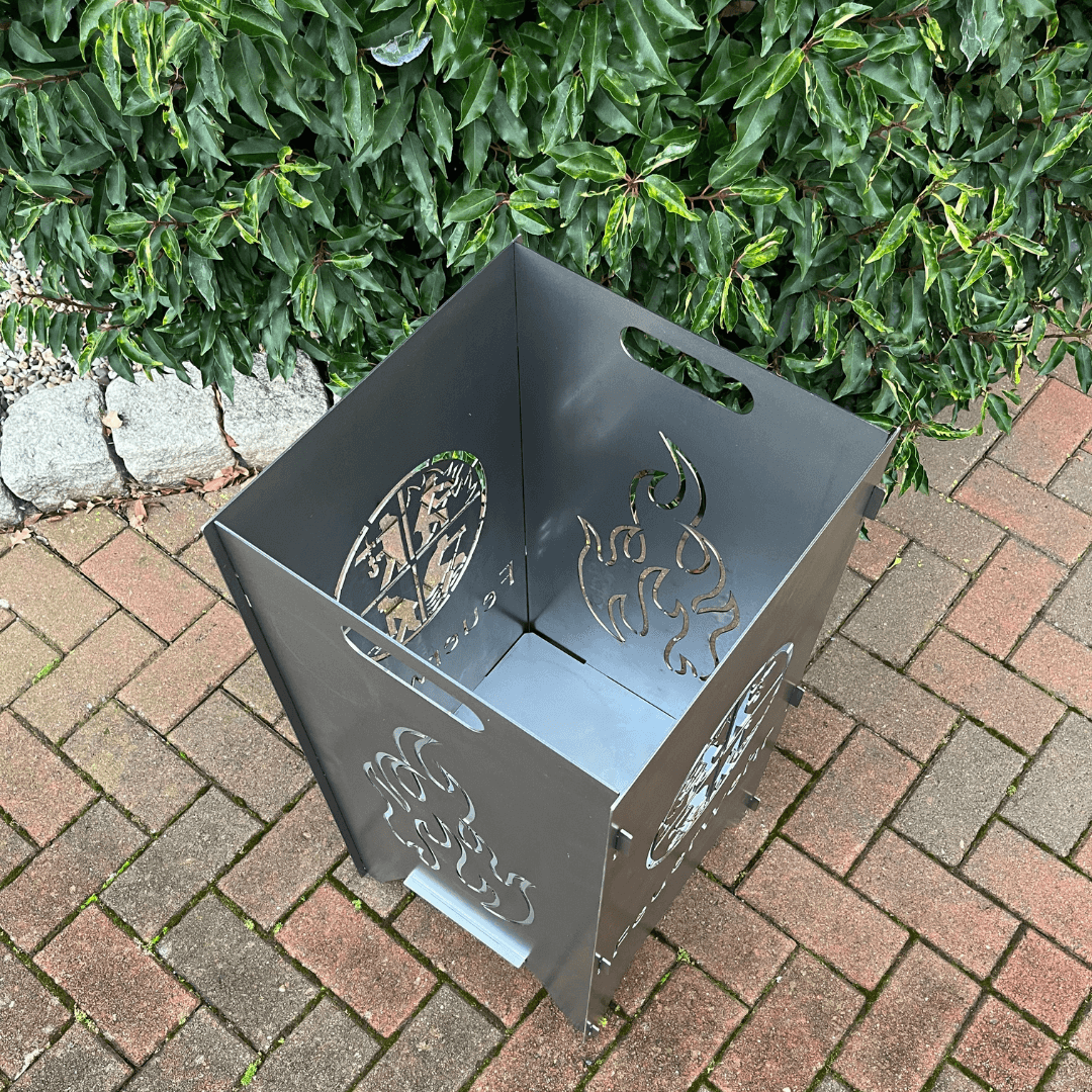 Feuertonne mit einem runden Feuerwehrwappen in der Mitte und darunter steht: "Feuerwehr" auf zwei Seiten. In dem runden Wappen sind Strichmännchen die verschiedenen Tätigkeiten nachgehen. Auf den anderen beiden Seiten befindet sich das Flammen-Motiv. Ansicht von oben.