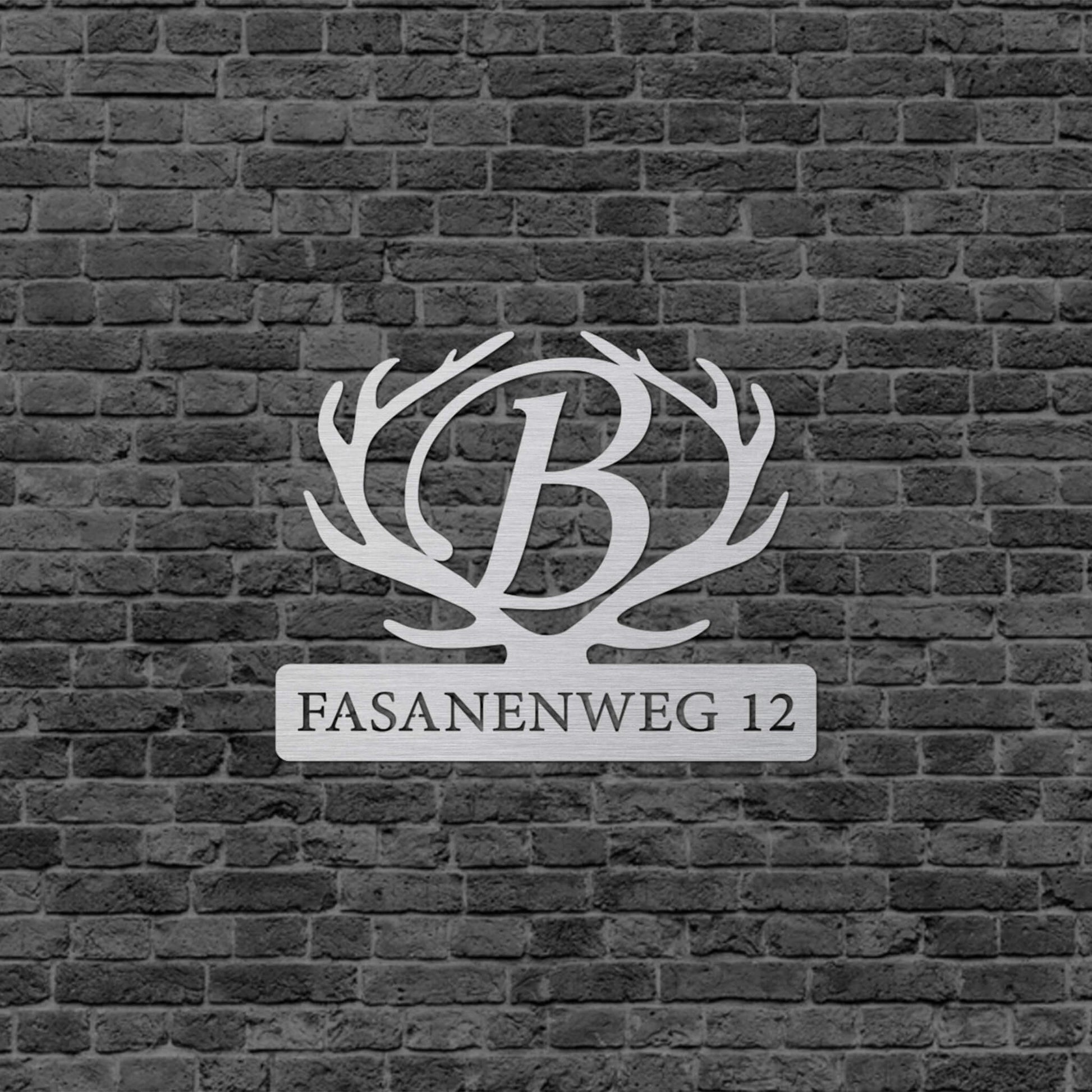 Eine Wanddekoration mit einem Hirschgeweih. In der Mitte des Hirschgeweihs befindet sich ein großes B und da drunter steht der Straßenname und die Nummer. Das Schild hängt an einer Hauswand.